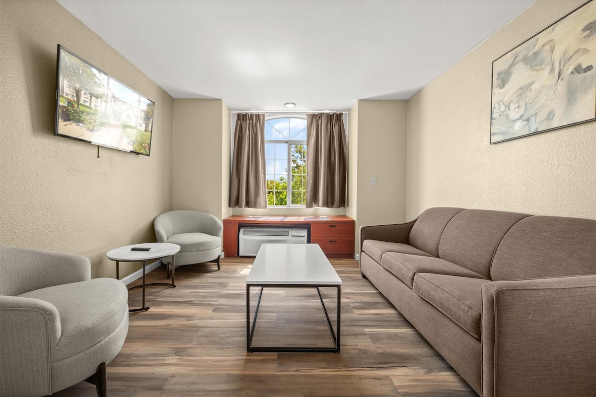 A living room with a sofa, armchair, and coffee table, a window with curtains, and a flat-screen TV mounted on the wall.
