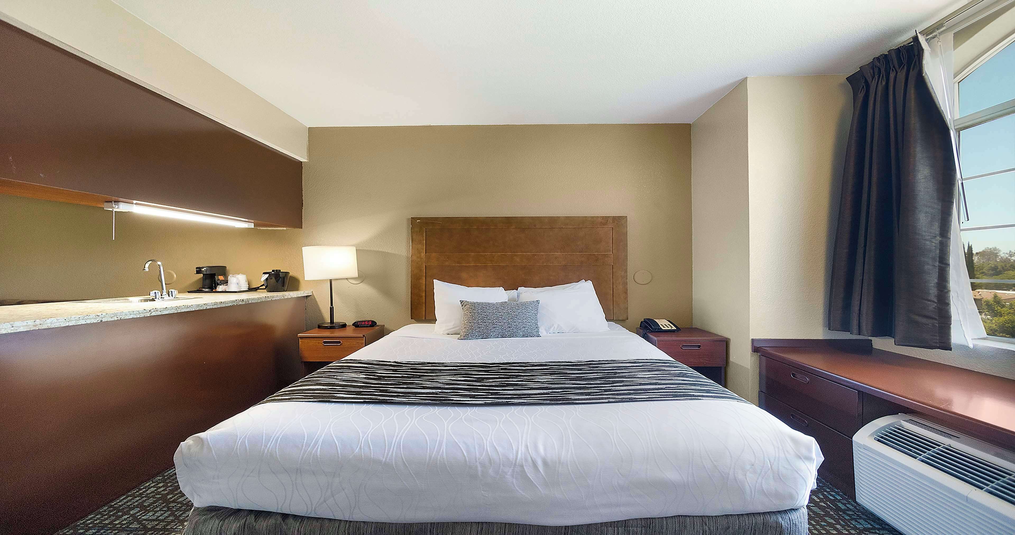 A hotel room with a large bed, a bedside table with a lamp, a phone, and a window with curtains.
