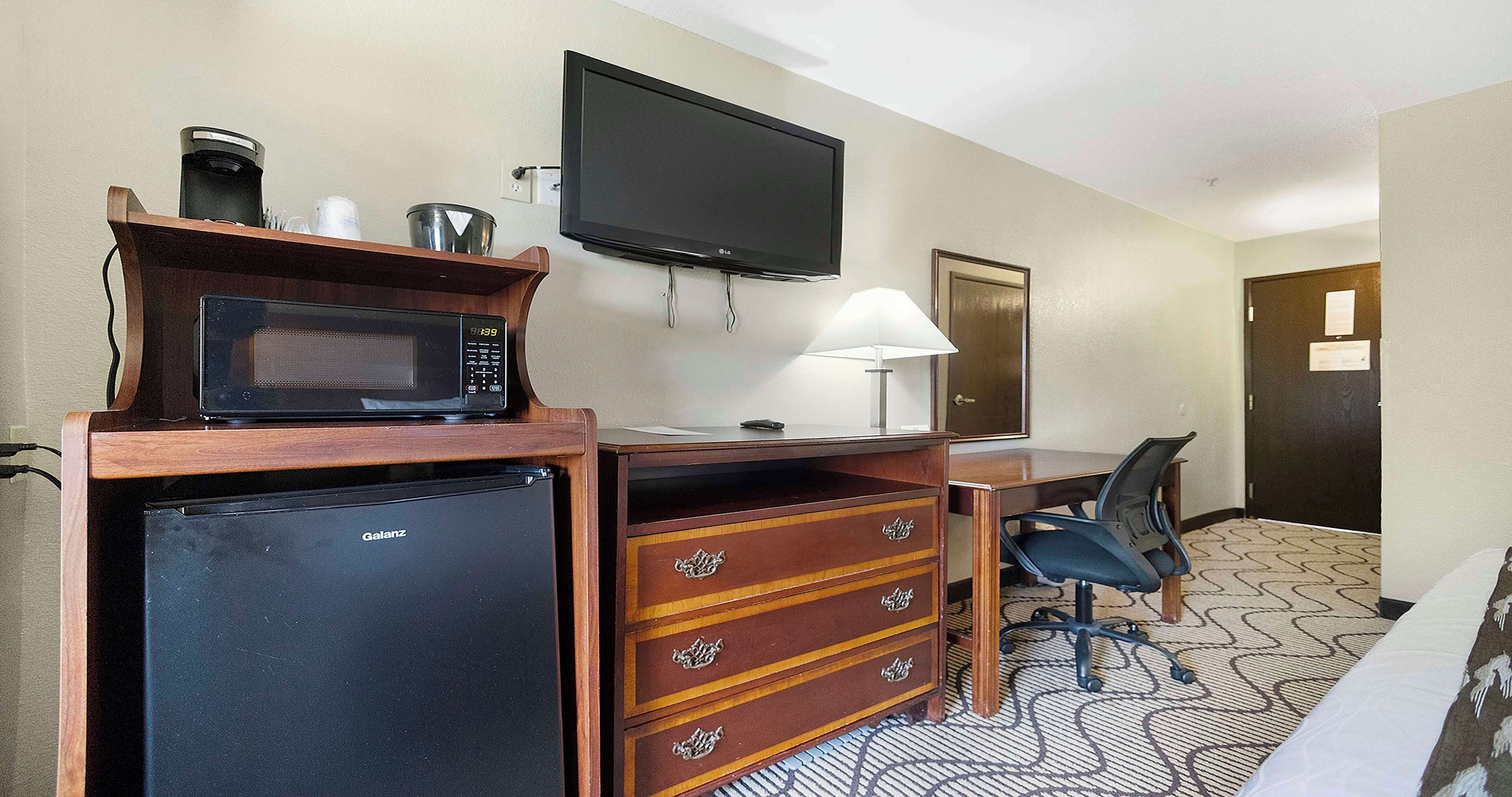 A hotel room with a minibar, a flat-screen TV, a desk with a lamp, a chair, and a bed.