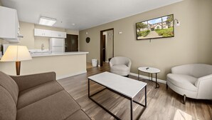 Un salon moderne avec un canapé, une table basse et deux fauteuils. On aperçoit, au fond, une cuisine avec des meubles blancs et un réfrigérateur.