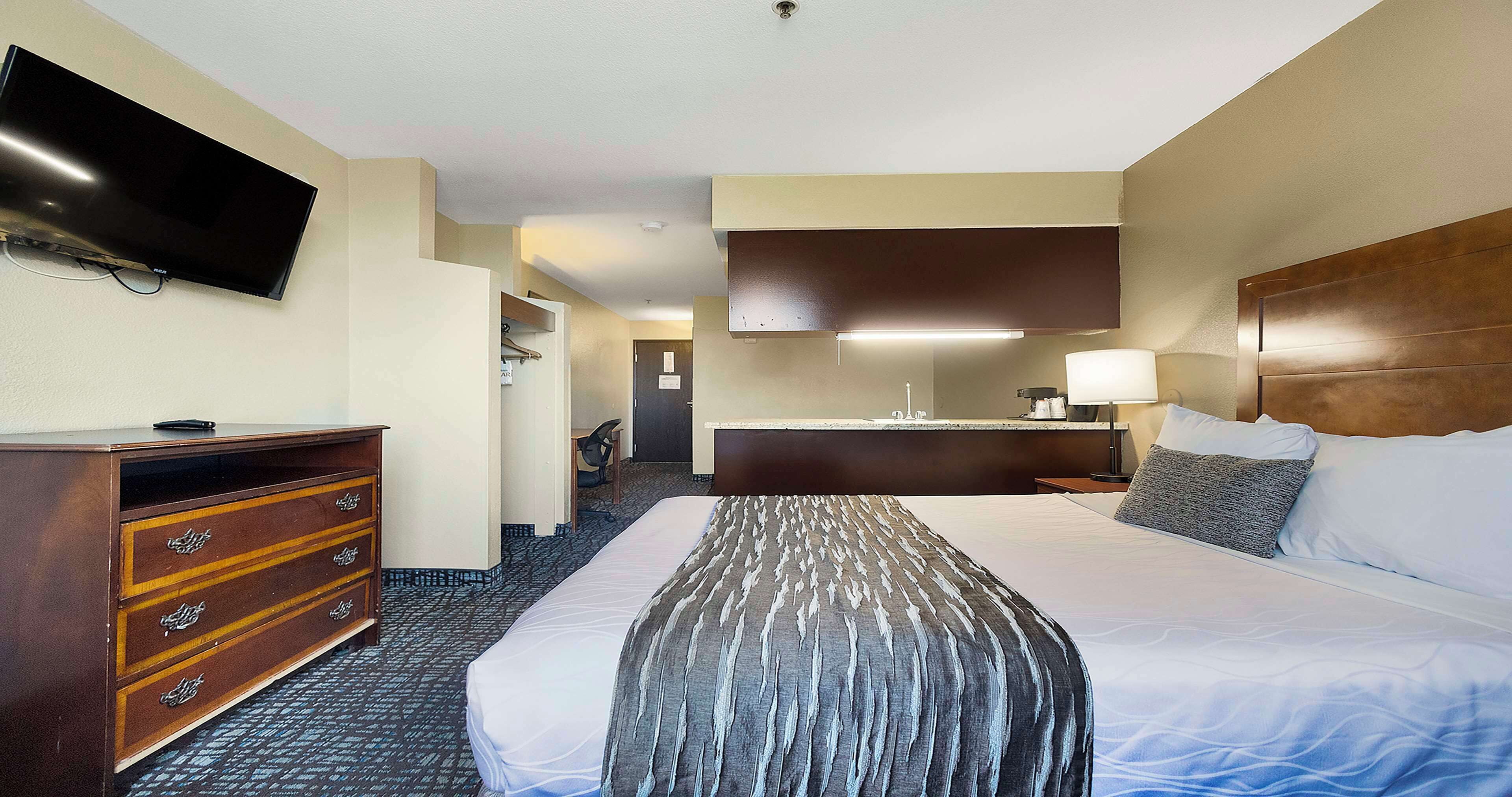 Een hotelkamer met een bed, een kaptafel, een aan de muur bevestigde televisie, een bureau met een stoel, en een badkamer die door een geopende deur zichtbaar is.