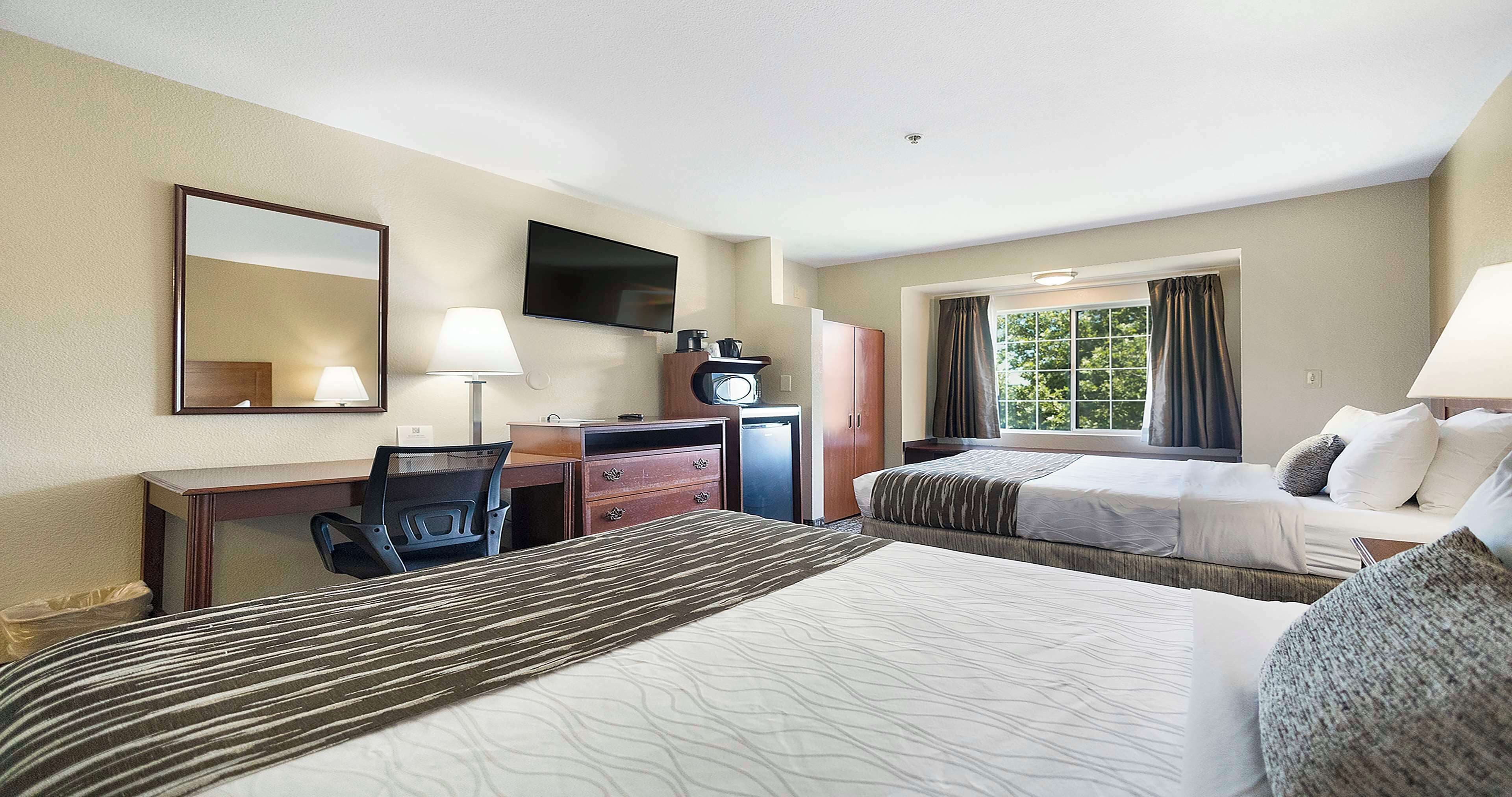 A hotel room with a large bed, a smaller bed, a desk with a chair, a TV mounted on the wall, and a window with curtains.