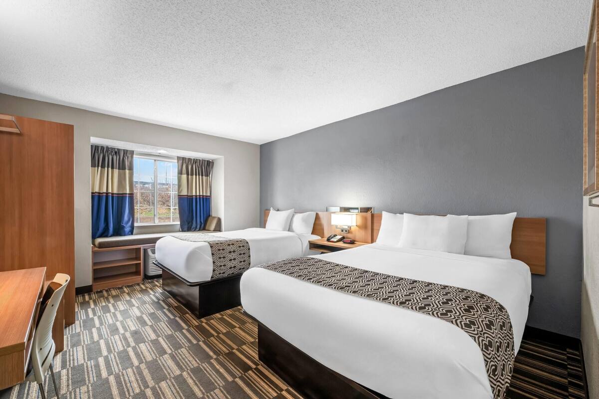 A hotel room with two beds, a desk, a chair, a window with curtains, and a lamp.