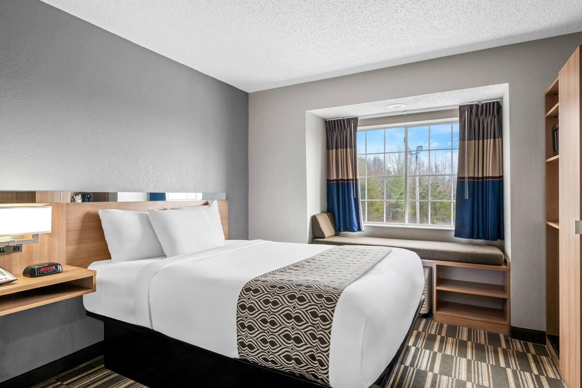 A hotel room with a large bed, a bedside table with a lamp, a window with curtains, and a built-in bench.