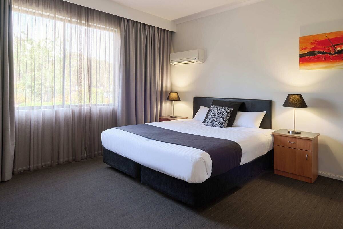 A hotel room with a large bed, two bedside tables, an air conditioning unit, and a window with curtains.