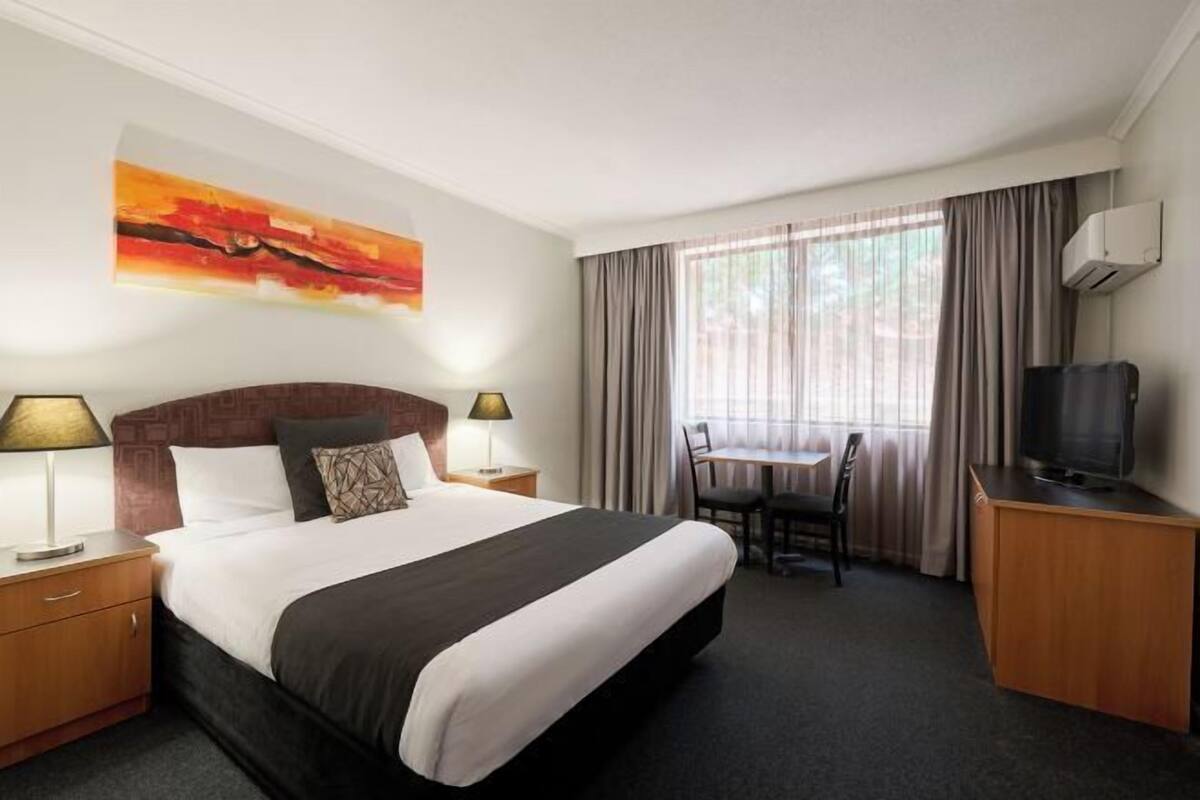 A hotel room with a bed, bedside lamps, a desk with a chair, a television, and a window with curtains.