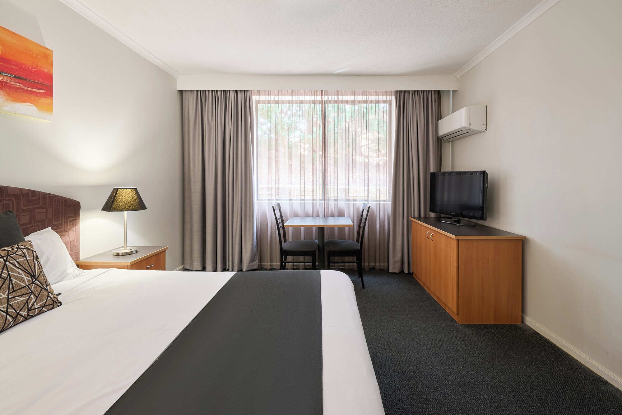 Quarto de hotel com uma cama, uma escrivaninha com televisão, uma cadeira, um abajur e uma janela com cortinas.