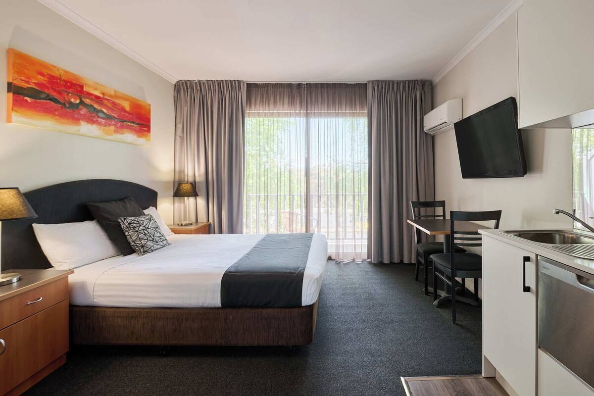 A hotel room with a bed, bedside table, lamp, TV, and a kitchenette.