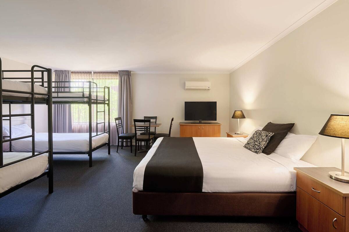 A hotel room with bunk beds, a desk, a TV, a lamp, and a bed with a black and white pillow.