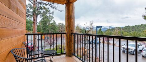 A wooden balcony with a table and chairs, overlooking a parking lot and a mountainous landscape.