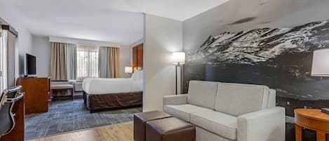 Ett hotellrum med en säng, en soffa, ett skrivbord, en stol, en tv och en stor tavla med ett bergsmotiv på väggen.