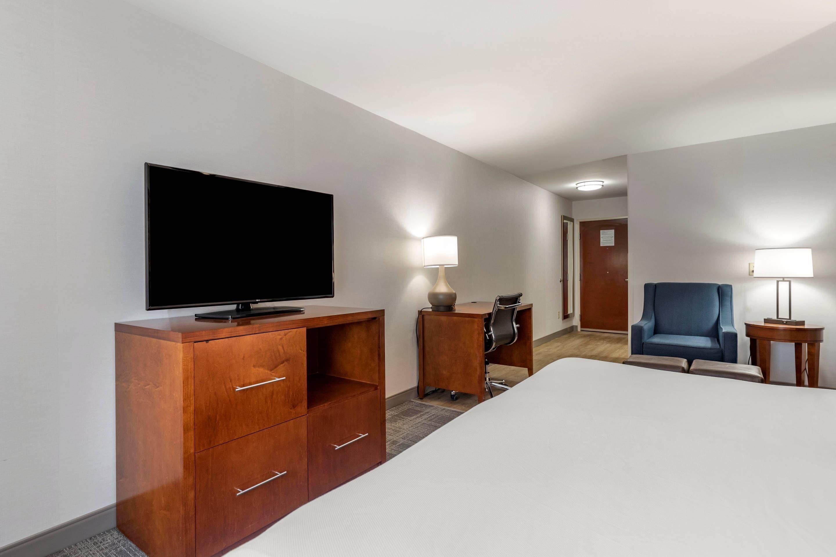 A hotel room with a bed, a TV, a desk with a chair, a lamp, and a blue armchair.