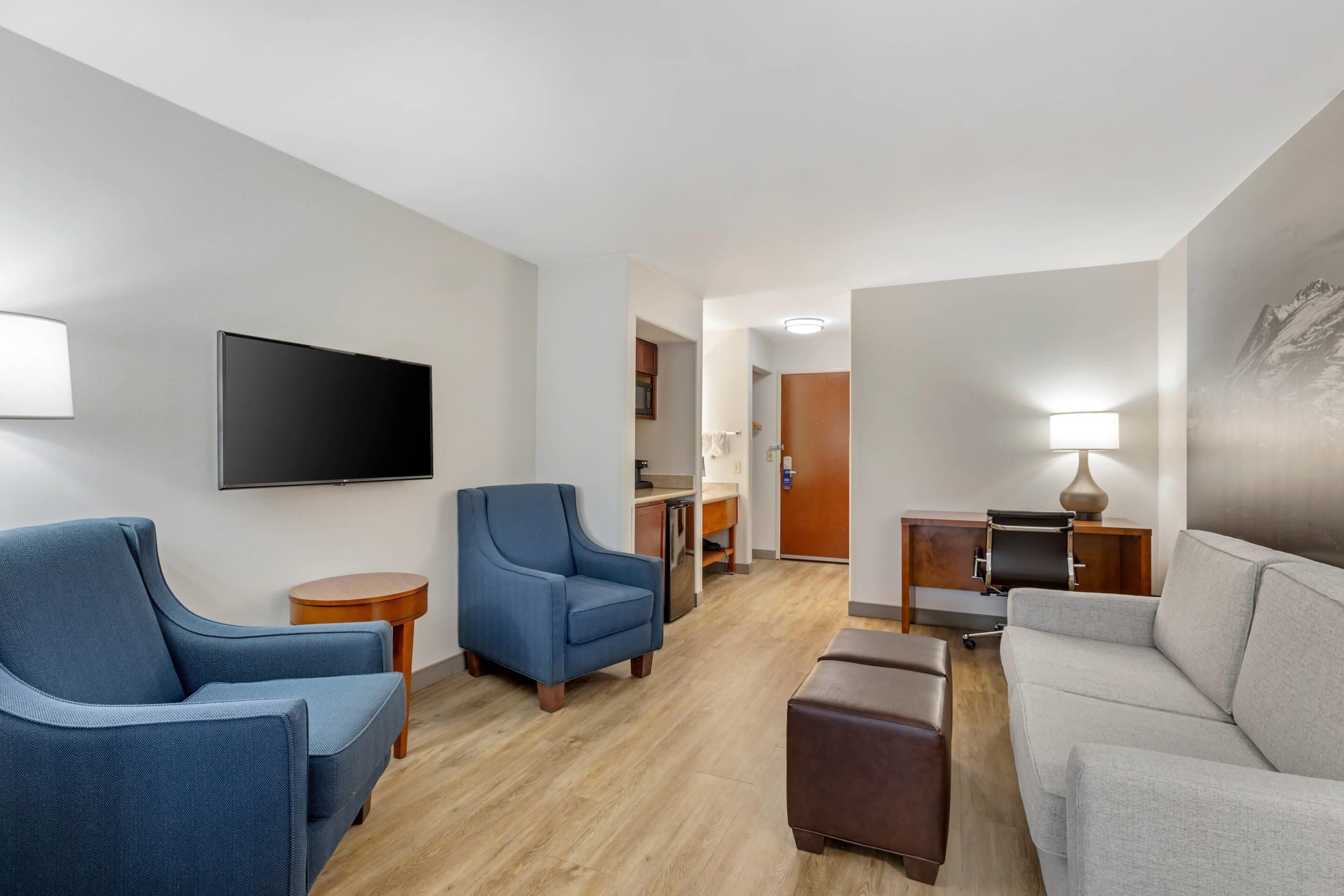 Ein modernes Wohnzimmer mit Flachbildfernseher, zwei blauen Sesseln, einem grauen Sofa, einem Holztisch und einem kleinen Beistelltisch mit Lampe.