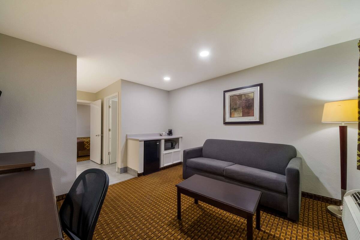 A hotel room with a sofa, a coffee table, a desk, a kitchenette, and a framed picture on the wall.