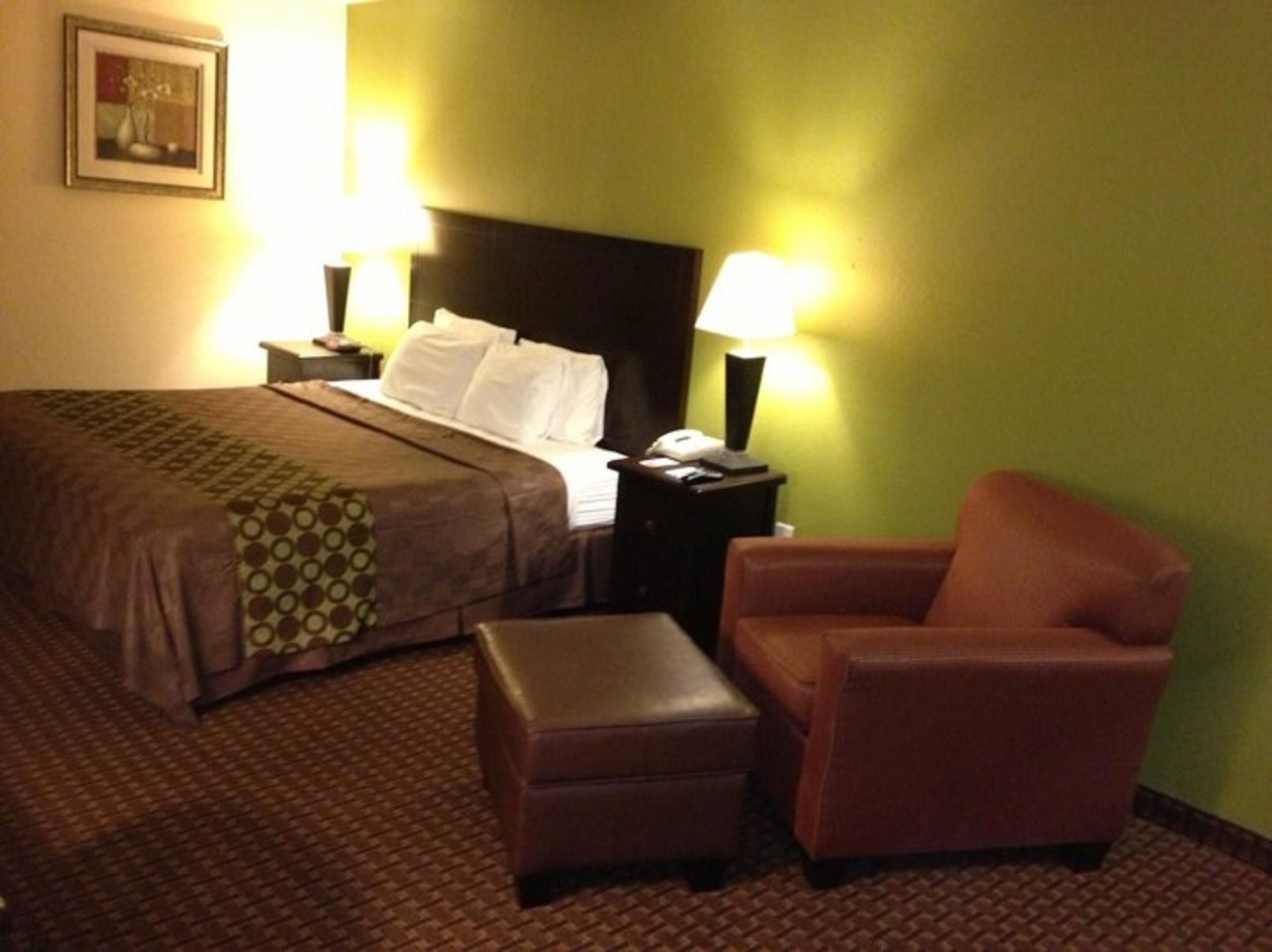 A hotel room with a bed, a chair, an ottoman, a nightstand, and a lamp.