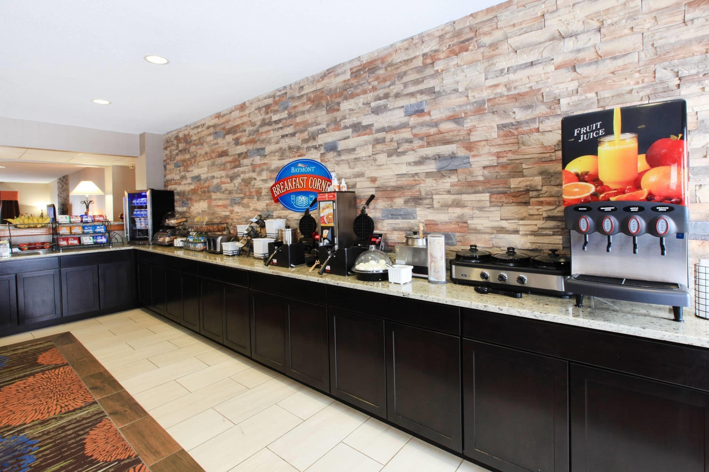 A breakfast bar with a sign for 'Baymont Breakfast Corner,' a coffee machine, a waffle maker, and a fruit juice dispenser.