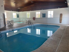 Una piscina interior con agua cristalina, rodeada de baldosas de piedra y equipada con sillas de piscina y un trampolín.