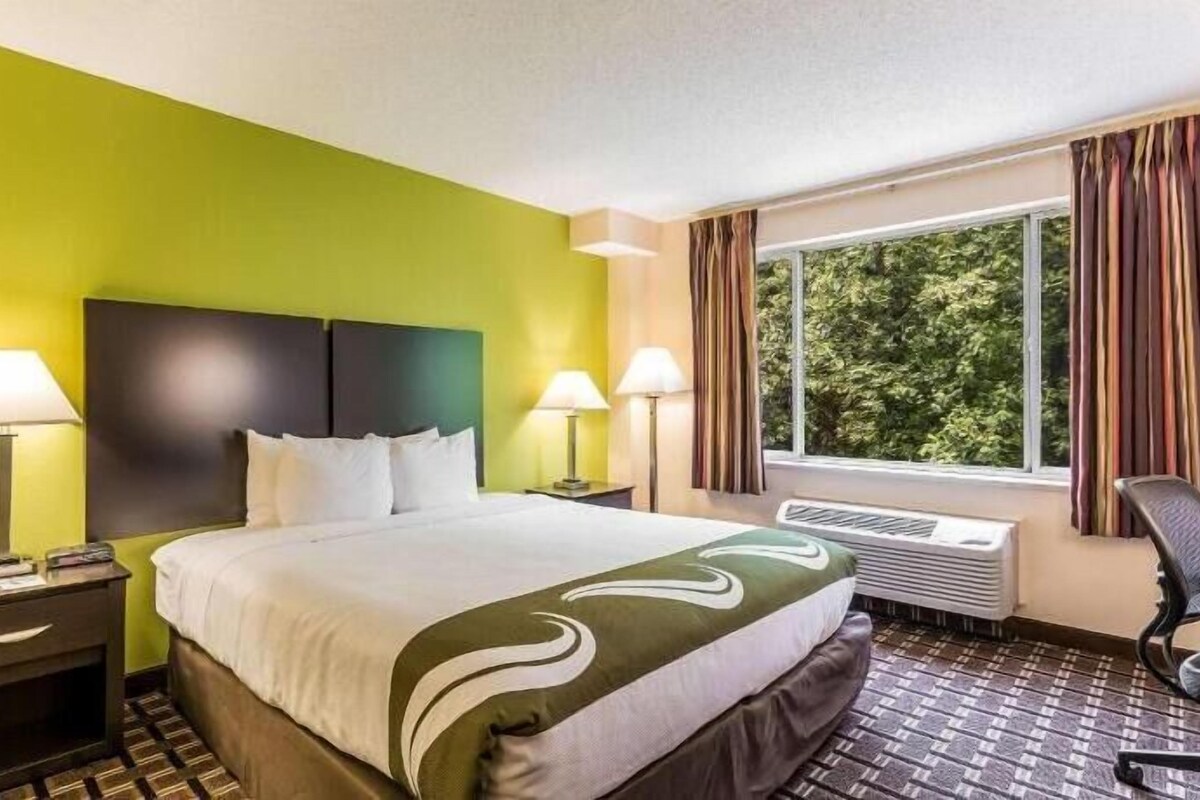 Une chambre d’hôtel avec un lit, deux lampes de chevet, une fenêtre avec des rideaux, une chaise et un radiateur.