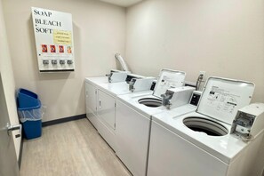 A laundry area with two washing machines, a soap and bleach dispenser, and a blue trash bin. - Comfort Inn Gallup I-40 Exit 20 (Gallup)