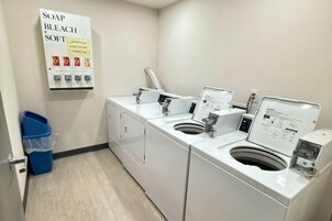 A laundry area with two washing machines, a soap and bleach dispenser, and a blue trash bin.