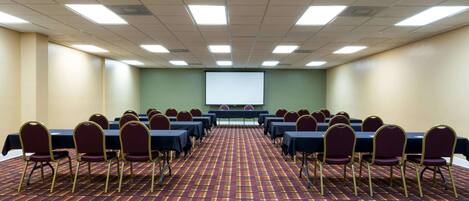 A conference room with rows of tables and chairs, a projector screen, and a patterned carpet.