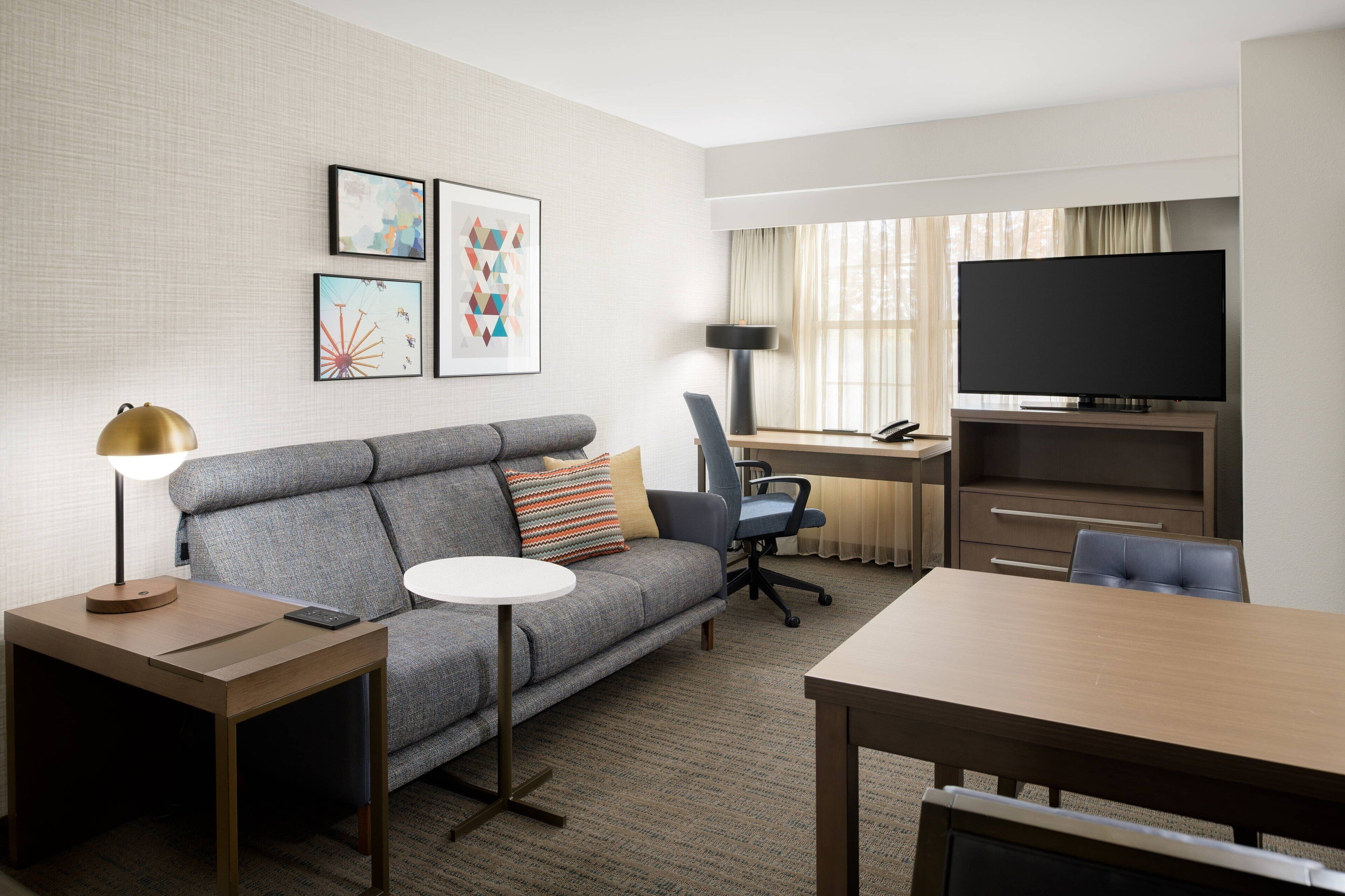 A hotel room with a grey sofa, a small round table, a desk with a chair, a television, and artwork on the wall.