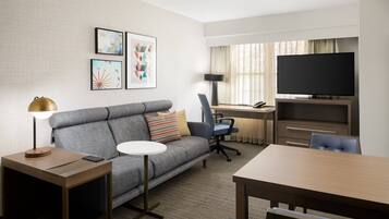 A hotel room with a grey sofa, a small round table, a desk with a chair, a television, and artwork on the wall.