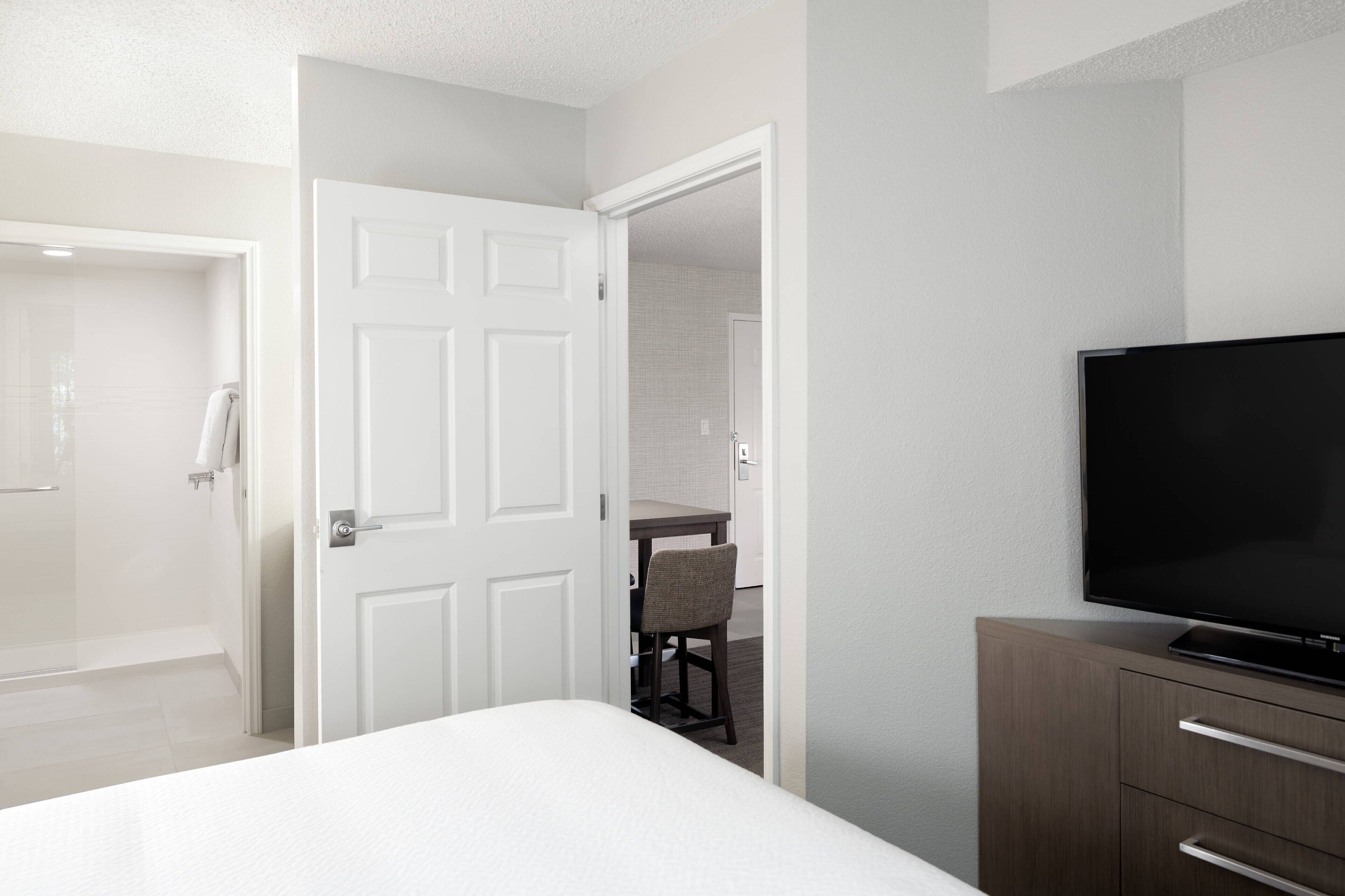 A hotel room with a bed, a TV, a wooden dresser, a bathroom with a shower, and a small dining area.