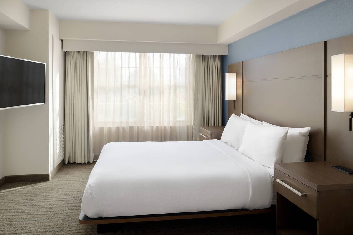 Habitación de hotel con una cama grande, una mesita de noche, un televisor de pantalla plana en la pared y una ventana con cortinas traslúcidas.