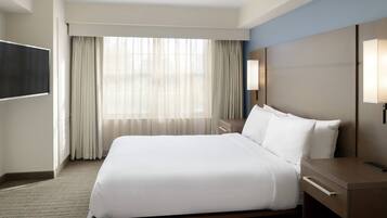 Habitación de hotel con una cama grande, una mesita de noche, un televisor de pantalla plana en la pared y una ventana con cortinas traslúcidas.