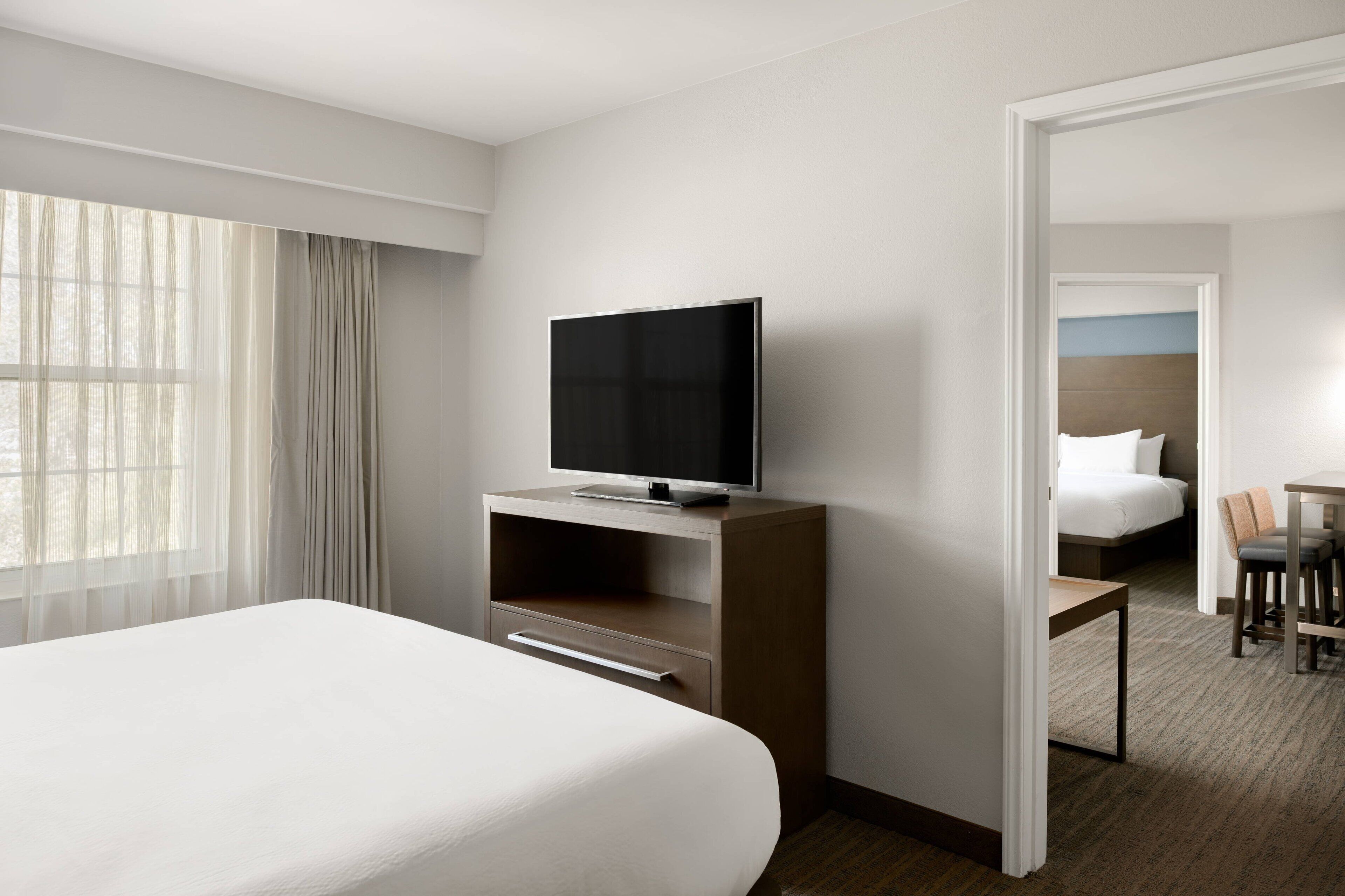 A hotel room with a large bed, a TV on a wooden stand, a desk with chairs, and a view of another room with a bed and a table.
