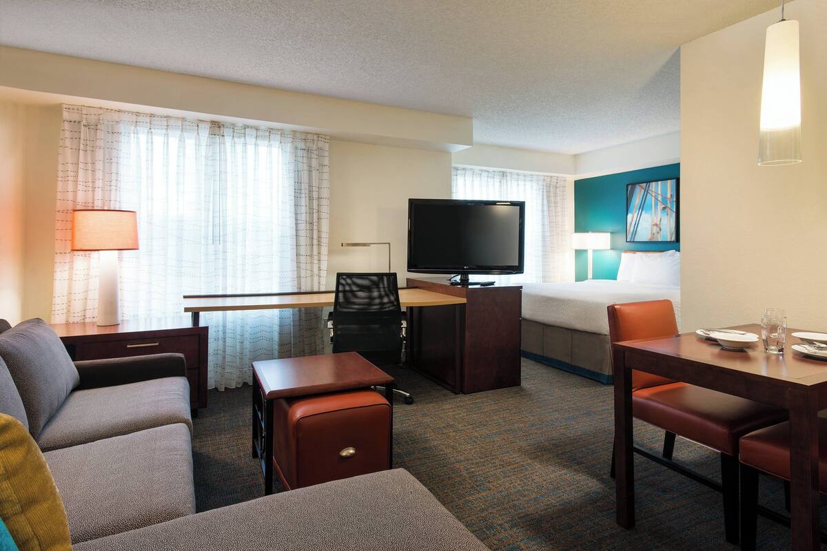Ein Hotelzimmer mit einer Couch, einem kleinen Tisch, einem Schreibtisch mit Fernseher, einem Bett und einem Essbereich mit Tisch und Stühlen.