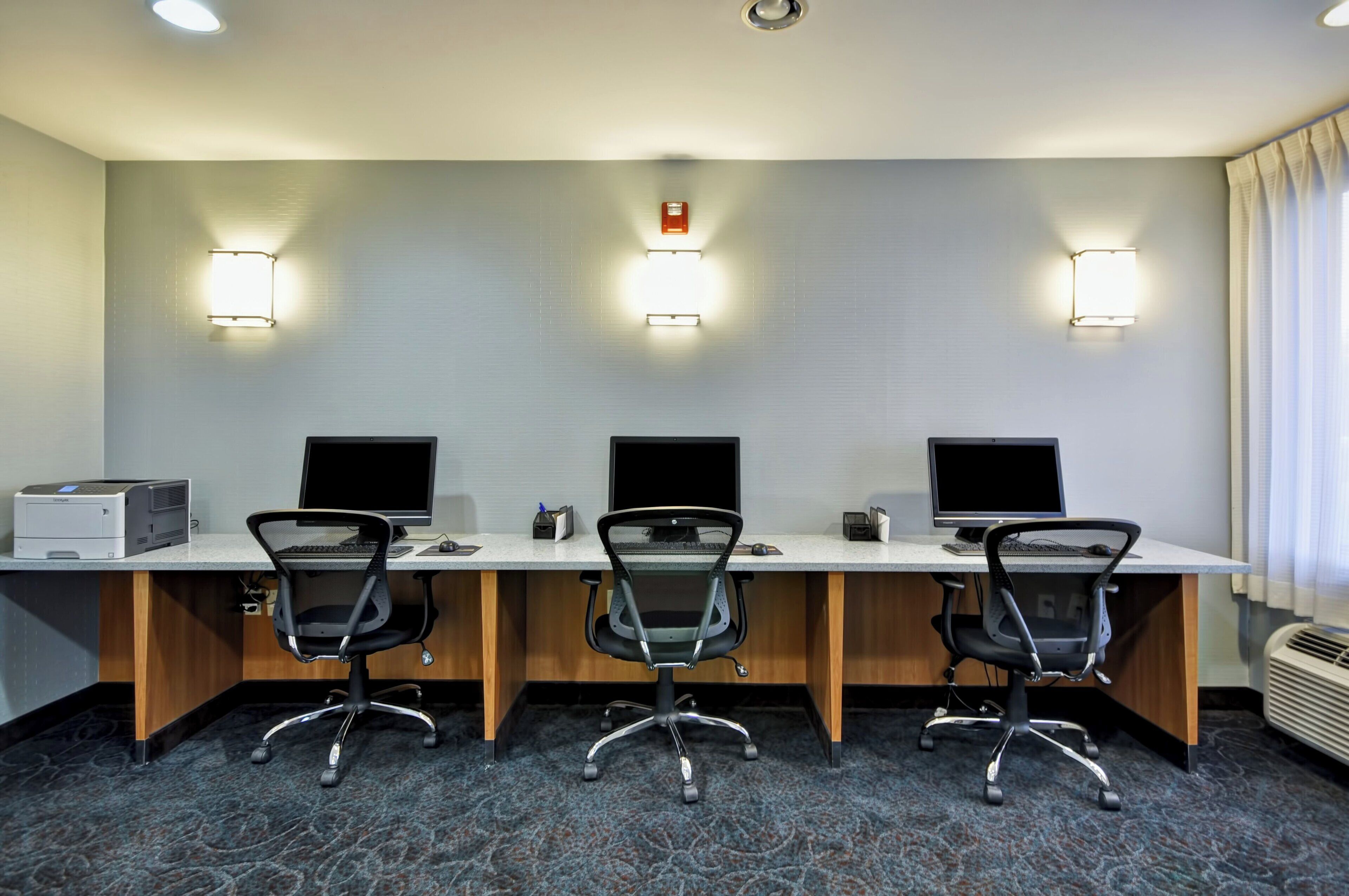 A hotel room with a desk, three chairs, and three monitors.