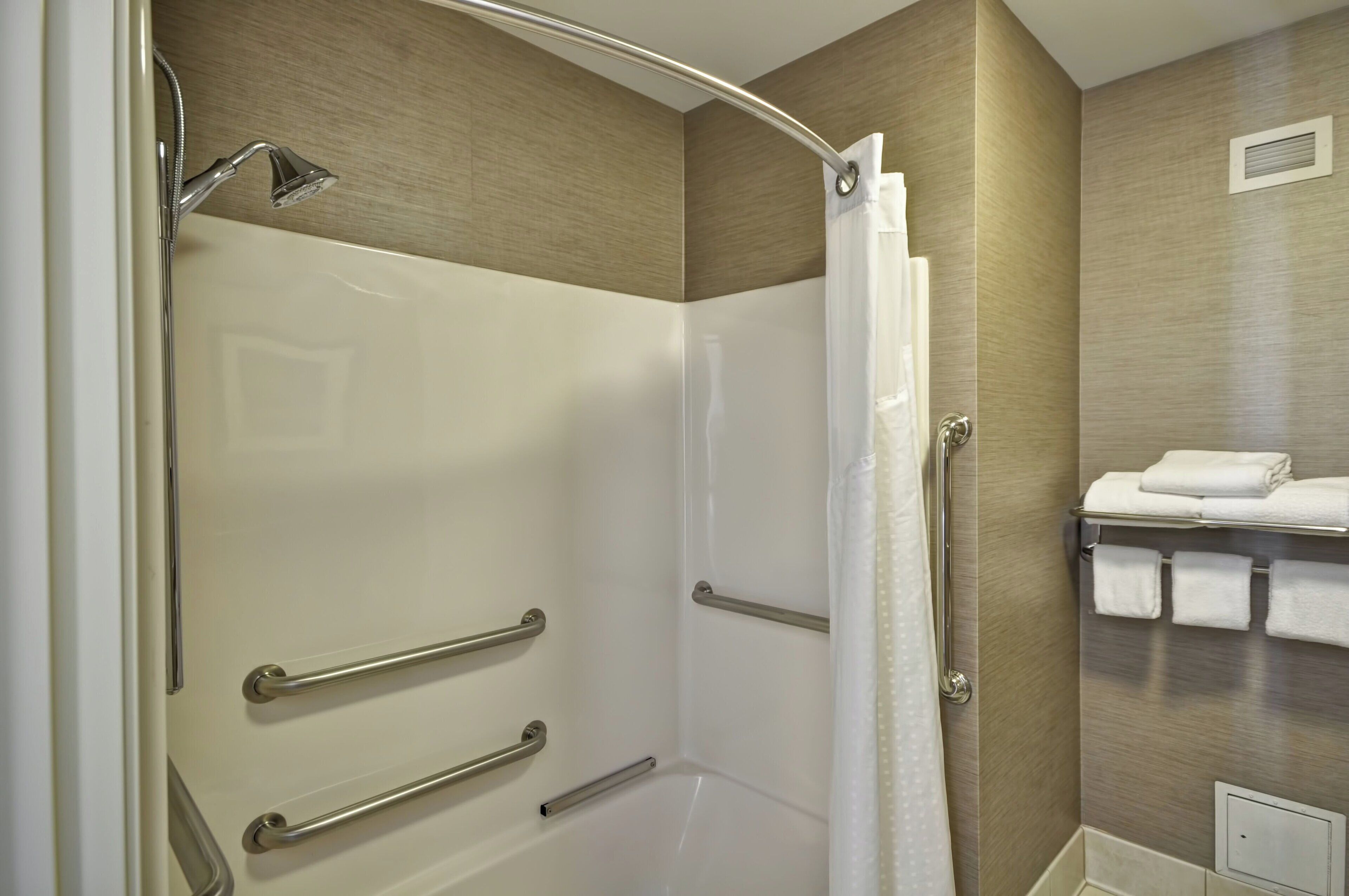 A modern bathroom with a walk-in shower, grab bars, a towel rack with neatly folded towels, and a wall-mounted vent.
