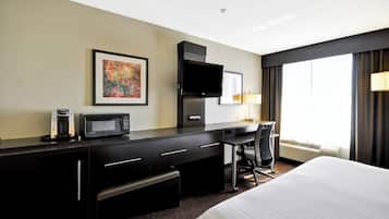 Habitación de hotel con cama, escritorio con televisor de pantalla plana, cafetera, microondas, silla y ventana grande con cortinas.