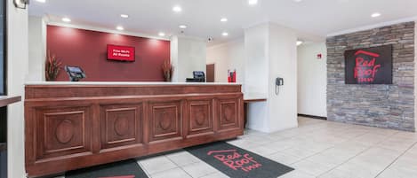 A hotel reception area with a wooden reception desk, a red wall with a 'Free Wi-Fi All Rooms' sign, a stone wall with a 'Red Roof Inn' sign, and a black and red doormat.