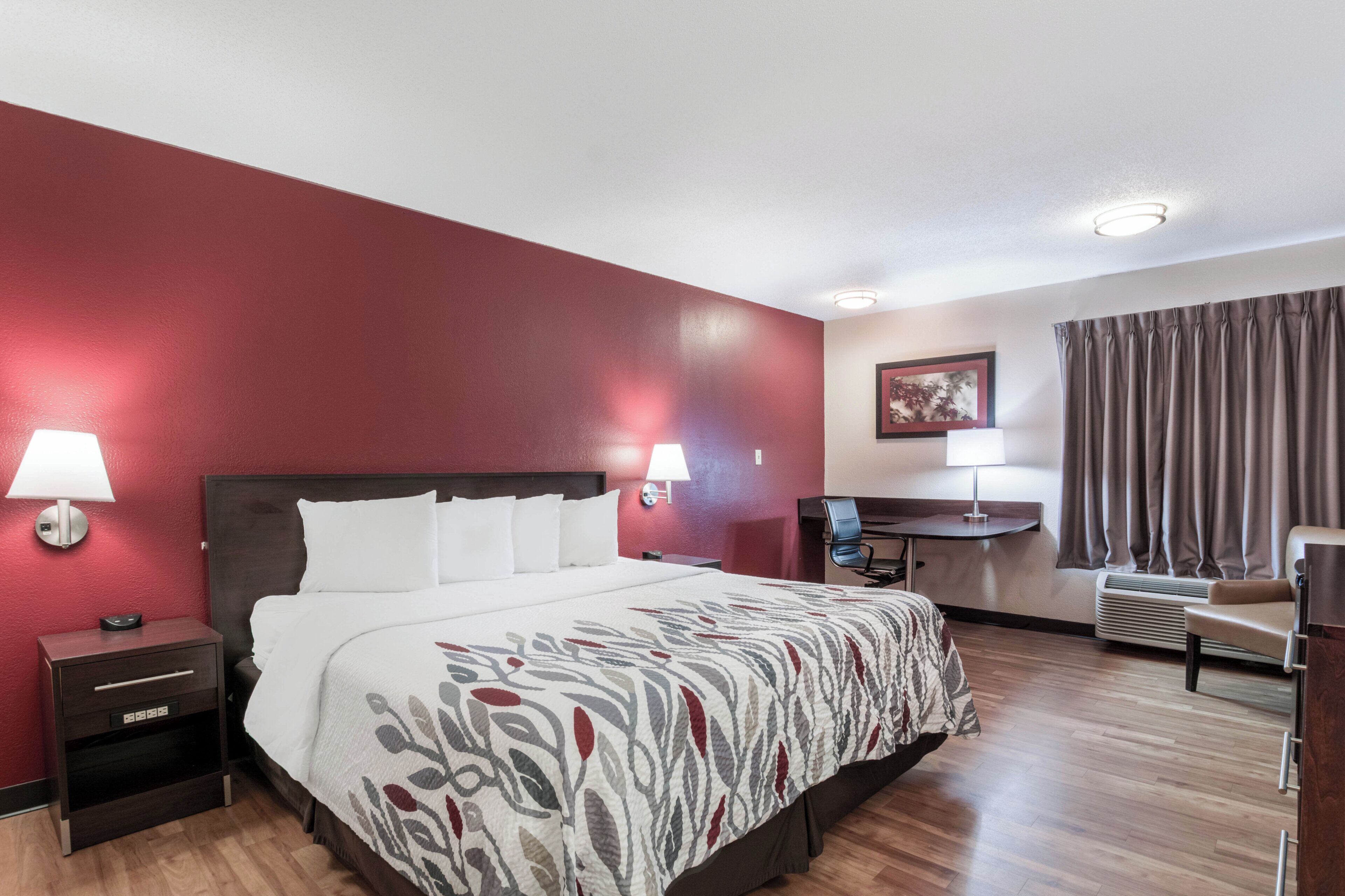Ein Hotelzimmer mit einem großen Bett, Nachttischlampen, einem Schreibtisch mit Stuhl, einem Fenster mit Vorhängen und einer Wand-Klimaanlage.