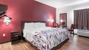 A hotel room with a bed, bedside table, lamp, chair, and an air conditioning unit.