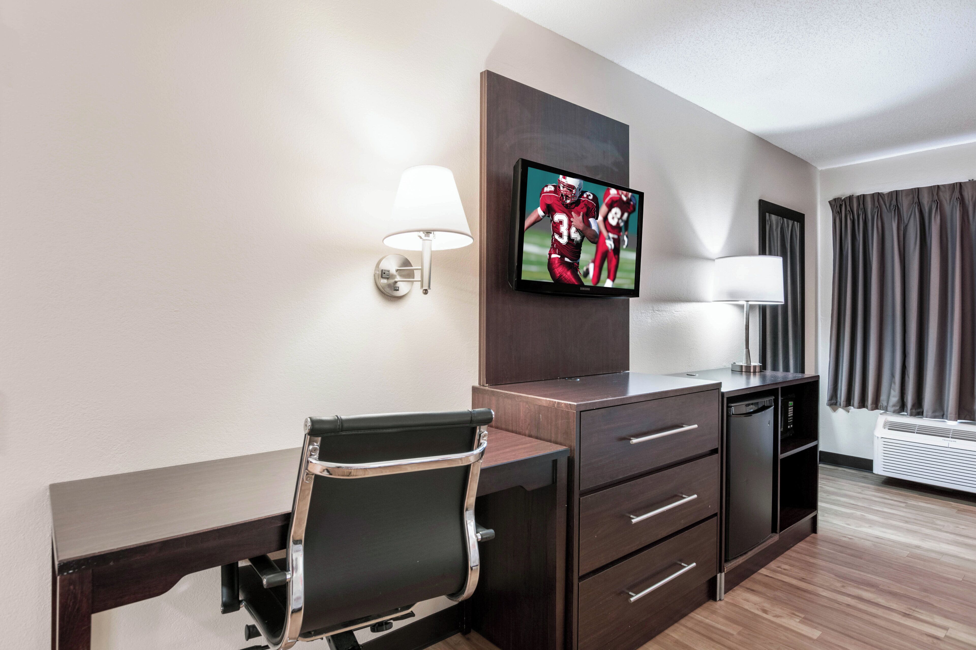 A hotel room with a desk, chair, TV, lamp, and air conditioning unit.