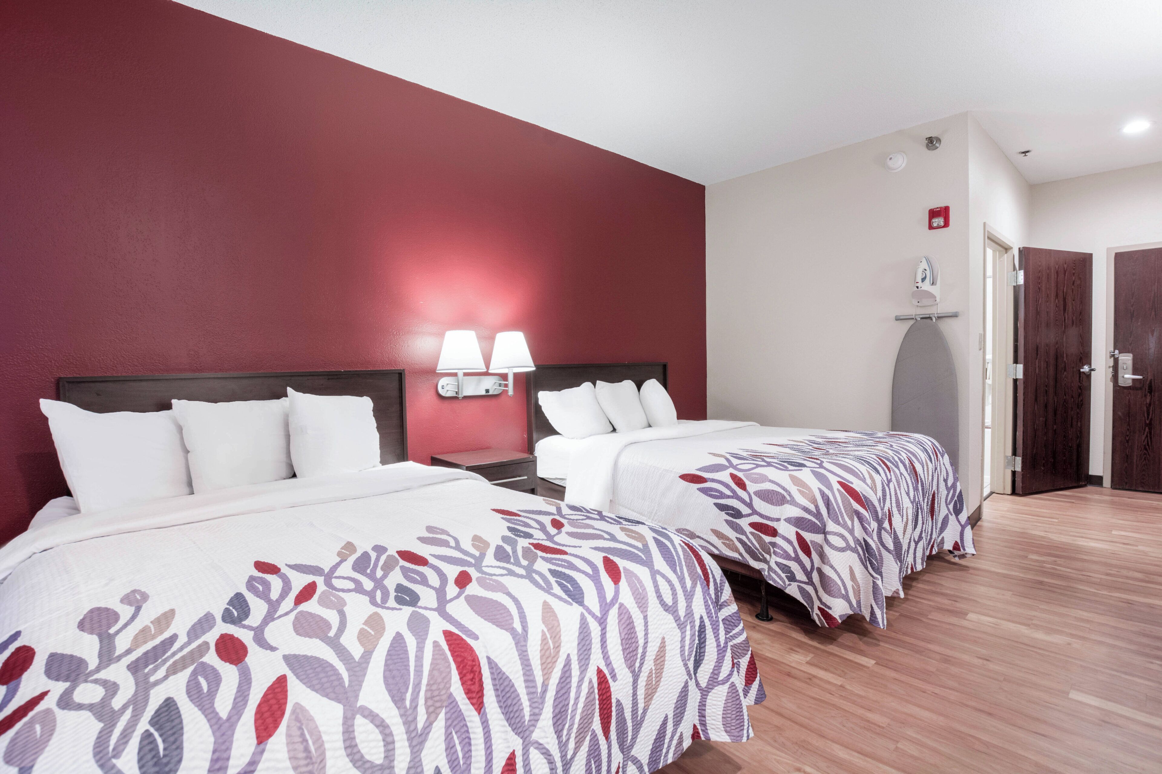 A hotel room with two beds, a red accent wall, a nightstand with lamps, and a door with a coat rack.