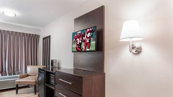 A hotel room with a flat-screen TV mounted on the wall, a desk with drawers, a lamp, a chair, and a window with curtains.