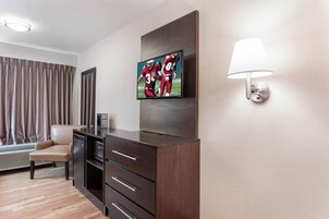 Habitación de hotel con televisor de pantalla plana montado en la pared, escritorio con cajones, lámpara, silla y ventana con cortinas.