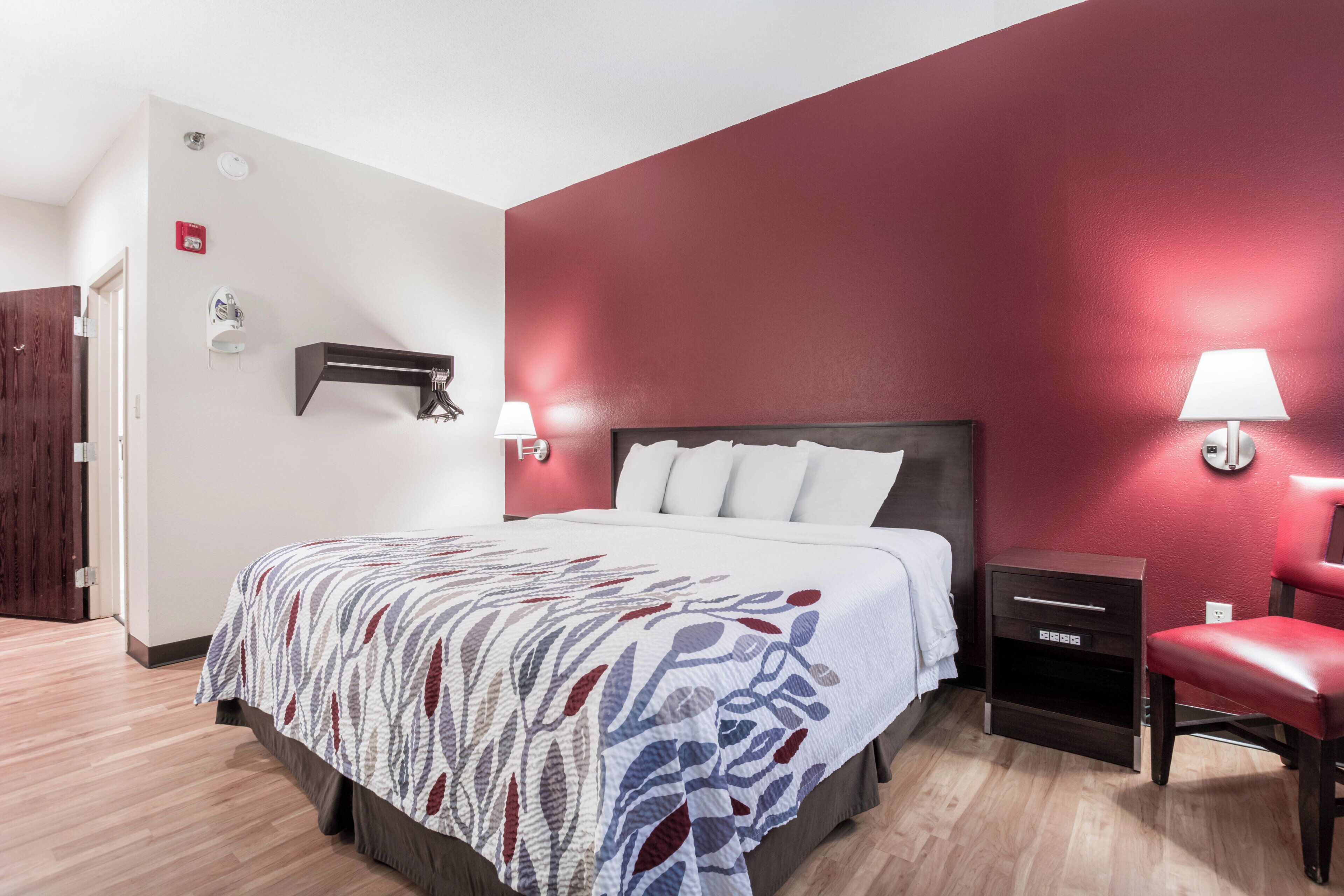 Habitación de hotel con cama, lámparas de noche, mesita de noche, una silla y una pared decorada en rojo.