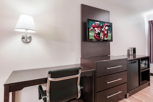 Habitación de hotel con televisión de pantalla plana montada en la pared, un escritorio con silla, una lámpara y una cafetera.