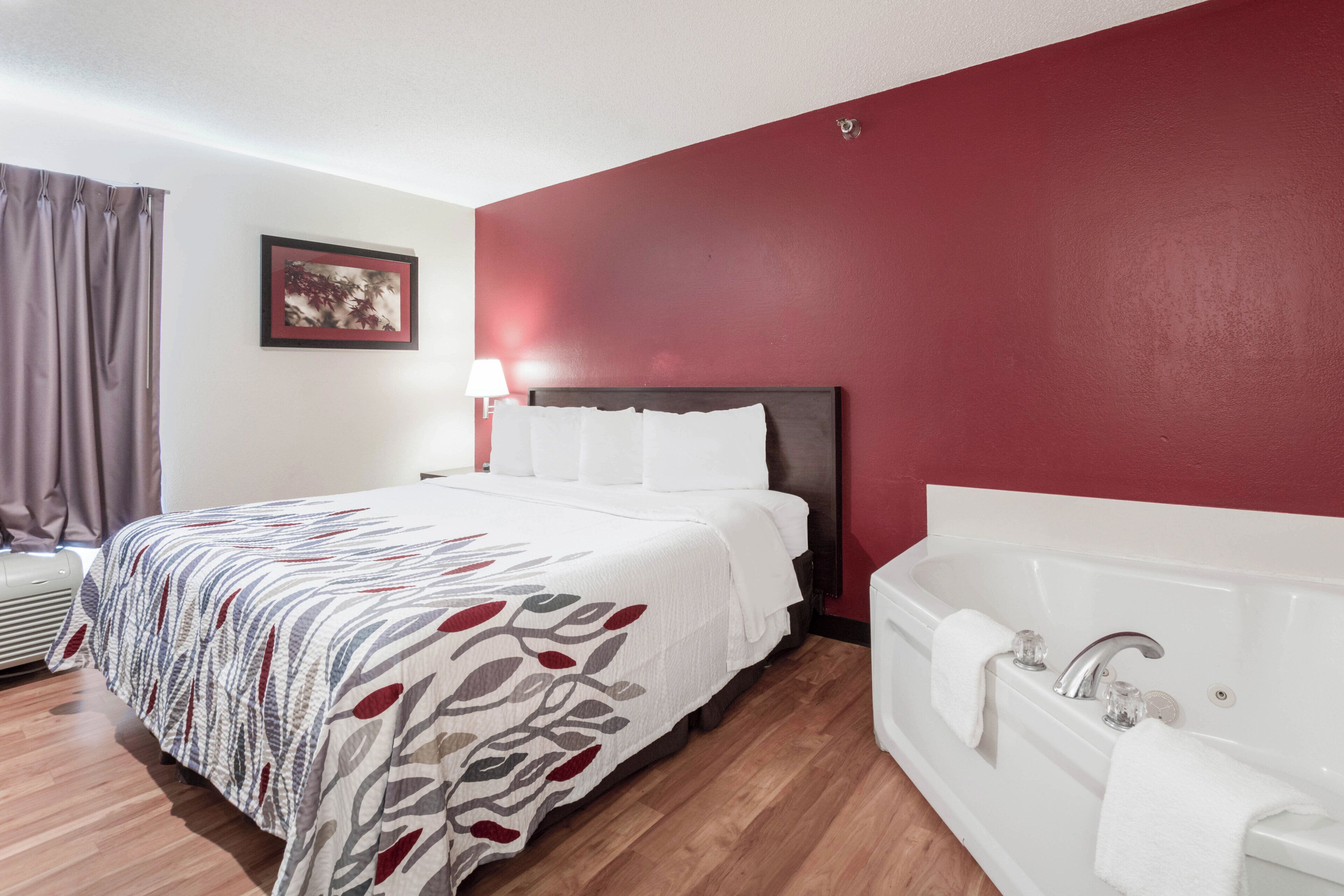 Habitación de hotel con cama, lámpara de noche, ventana grande con cortinas, un cuadro en la pared y un baño independiente con bañera.