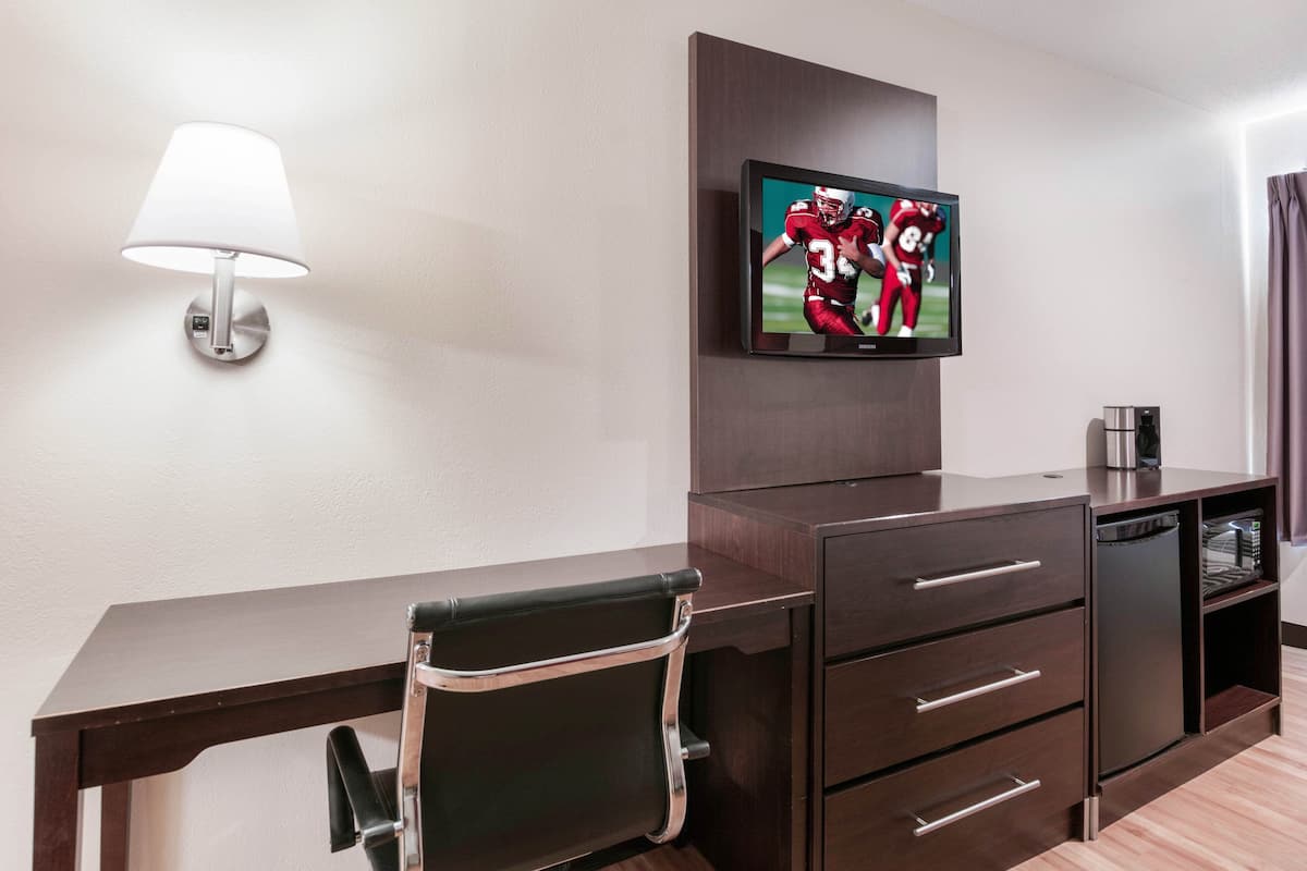 A hotel room with a desk, a chair, a TV mounted on the wall, a lamp, and a coffee maker.