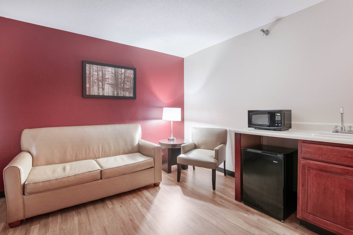 A compact living space with a beige sofa, a small table with a lamp, a microwave, and a compact refrigerator.