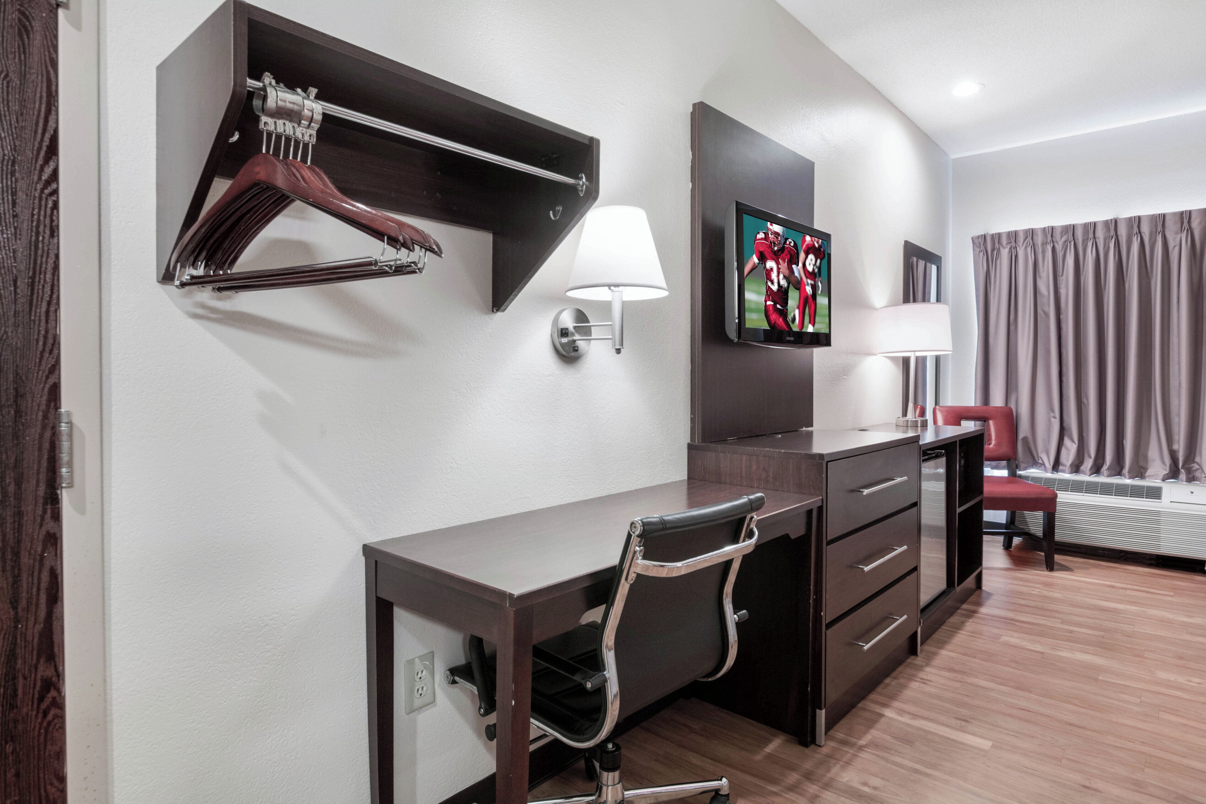 Habitación de hotel con escritorio, silla, perchero, televisor montado en la pared y una silla roja.