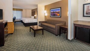 Una habitación de hotel con cama, sofá, mesa de centro, televisión, escritorio y una pequeña cocina.