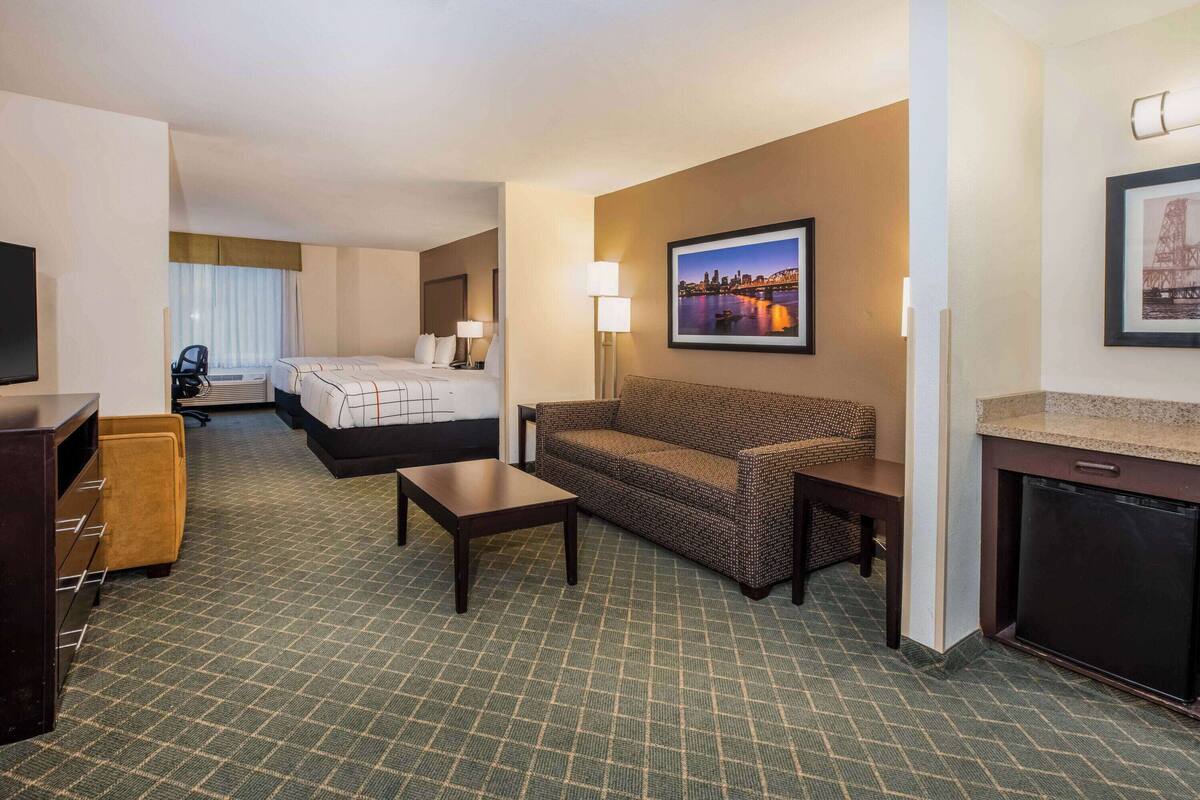 Una habitación de hotel con cama, sofá, mesa de centro, televisión, escritorio y una pequeña cocina.